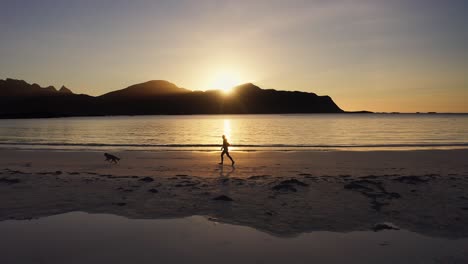 Lateral-tracking-shot-of-a-jogger-running-on-a-beach-with-her-golden-retriever-dog-at-sunset,-Lofoten-Islands,-Norway