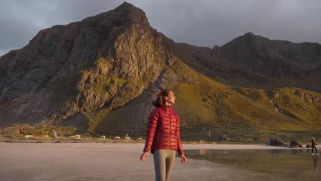 SLOW-MOTION,-a-beautiful-young-woman-in-a-red-jacket-admiring-the-view-and-smiling-on-the-Flakstad-beach-in-front-of-a-gorgeous-golden-sunset,-Lofoten-Islands,-Norway