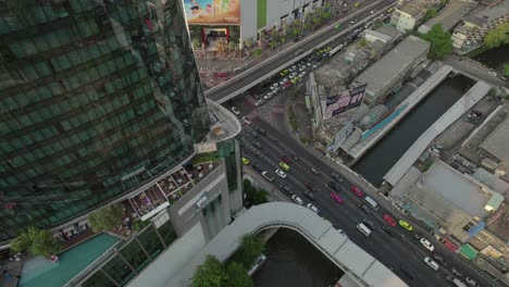 Aerial-of-the-vibrant-road-traffic-bustling-near-Bangkok's-towering-skyscrapers,-capturing-the-dynamic-essence-of-urban-life-in-Thailand's-capital