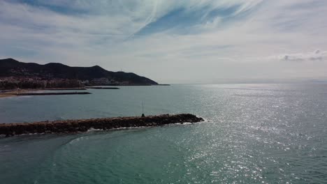 Sitges-coast-with-sparkling-sea-and-mountainous-horizon-on-a-sunny-day,-aerial-view
