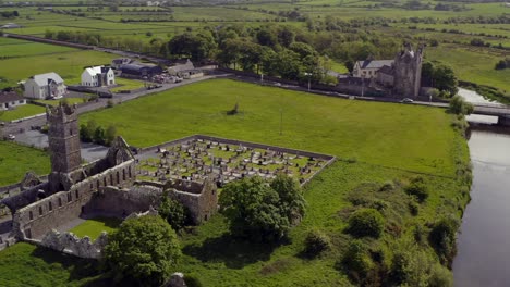 Claregalway-Friary-ruins-and-cemetery-opposite-the-castle,-aerial-orbit-on-sunny-day-in-Ireland