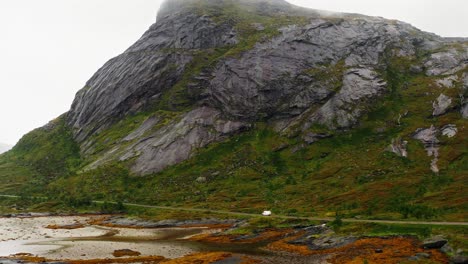 Vista-Aérea-De-Una-Autocaravana-Blanca-En-Una-Carretera-Costera-Al-Pie-De-Una-Montaña-Majestuosa,-Islas-Lofoten,-Noruega