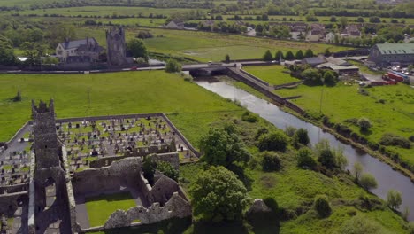 Claregalway-Friary-ruins-and-castle-on-sunny-vibrant-day,-aerial-by-river-Clare