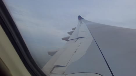 Blick-Durch-Das-Flugzeugfenster-Auf-Die-Tragfläche-Eines-Flugzeugs-Mit-Sich-öffnenden-Klappen,-Das-Durch-Wunderschöne-Wolken-Fliegt