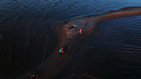 Drone-circling-kids-building-sand-castles-on-a-sandbank-in-shallow-water,-sunset