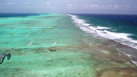 A-vibrant-coral-reef-with-kite-surfers-and-boats,-clear-blue-waters-under-a-sunny-sky,-aerial-view