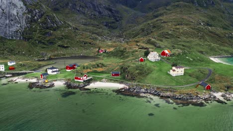 Vista-Aérea,-órbita-Inclinada-Hacia-Arriba,-Algunas-Casas-De-Rorbu-Rojo-En-Un-Pequeño-Pueblo-Pesquero-Costero-Al-Pie-De-Majestuosas-Montañas-En-Las-Islas-Lofoten,-Noruega