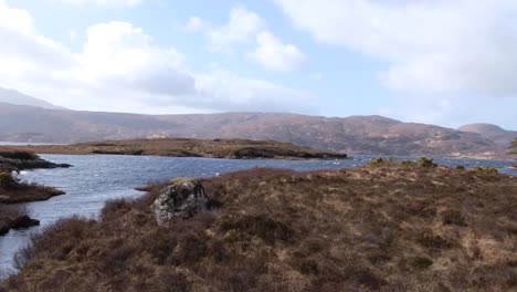 Slow-pan-of-Lewisian-gneiss-landscape-overlooking-Loch-Inver-water,-islands-with-trees,-and-tussock-grasses-in-the-highlands-of-Scotland-UK