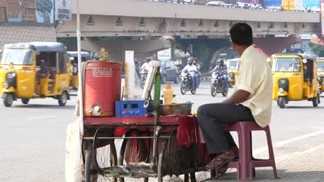 A-man-sits-at-a-drink-stand-beside-a-road-in-India,-Hyderabad-as-vehicles-drive-past