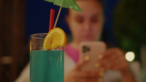 Woman-using-smartphone-sits-in-front-of-blue-drink-with-straw-and-umbrella