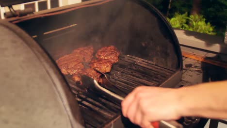 La-Mano-Masculina-Caucásica-Mueve-Filetes-Gruesos-Y-Jugosos-Cocinando-Y-Fumando-En-Una-Parrilla-Al-Aire-Libre-En-El-Patio-Trasero,-Cerca-De-La-Sartén