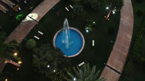 aerial-shot-of-a-small-pool-fountain-in-a-greenish-park-with-lights-on-the-grass