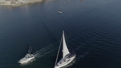 Aerial-shot-of-a-white-sailboat-and-a-fishing-boat-heading-into-the-Newport-Bay-in-Newport-Beach,-California-with-Pirates-Cove-in-the-background