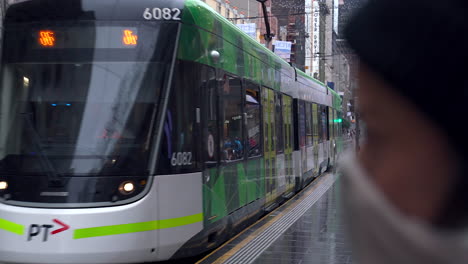 Los-Tranvías-De-Melbourne-Pasan-Esperando-A-Un-Viajero-Con-Una-Máscara-Facial,-En-El-Centro-Húmedo-De-La-Ciudad.