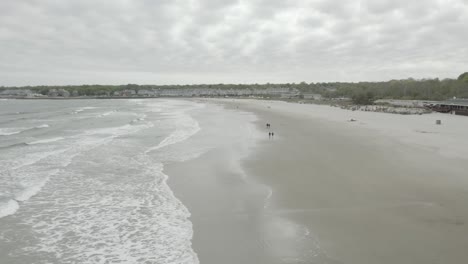 Drone-flying-to-the-right-and-panning-to-the-left,-while-people-enjoy-a-walk-on-the-beach,-cloudy-weather