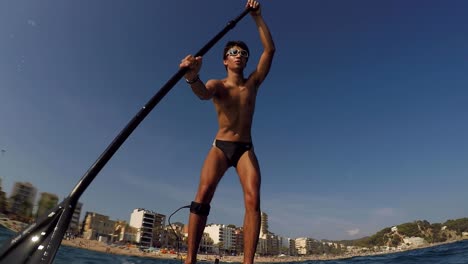 Young-man-in-swimsuit,-standup-paddling-with-city-beach-in-the-background