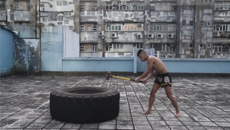 Ein-Mann-Macht-Übungen-Auf-Dem-Dach-Eines-Gebäudes-In-Hongkong,-Während-Er-Mit-Einem-Schweren-Hammer-Auf-Einen-Großen-Reifen-Schlägt