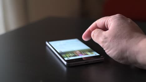 Close-up-male-hand-using-smartphone-scrolling-through-Facebook-on-table
