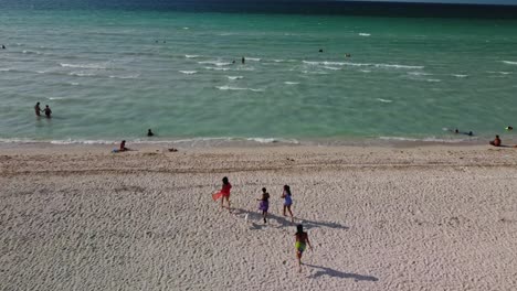 A-group-of-friends-running-on-the-sand-beach,-tourists-swimming-in-the-clear-water,-and-frothy-tides-can-be-observed-in-the-top-view-taken-by-a-drone