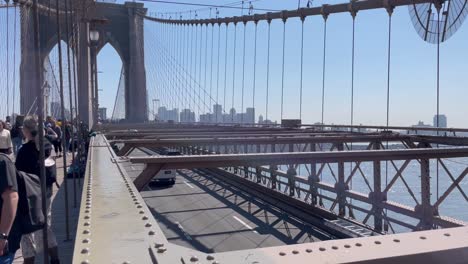 Schwenkaufnahme-Von-Touristen,-Die-Einen-Frühlingstag-Auf-Der-Brooklyn-Bridge-In-New-York-City-Genießen,-Mit-New-Jersey-Im-Hintergrund-Und-Darunter-Vorbeifahrenden-Fahrzeugen