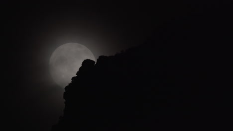 A-full-moon-rises-behind-clouds-and-a-rocky-mountain-foreground