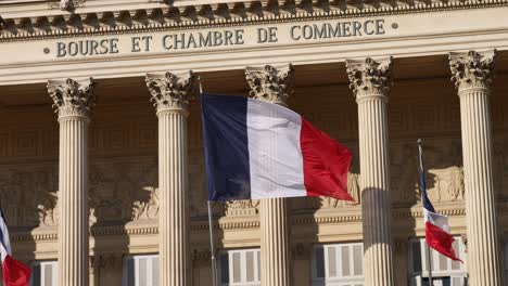 Bandera-Francesa-Moviéndose-En-El-Viento-Frente-A-Un-Edificio-Inspirado-En-Las-Antiguas-Columnas-Griegas-Con-Su-Sombra-En-La-Pared-Del-Centro-De-Negocios