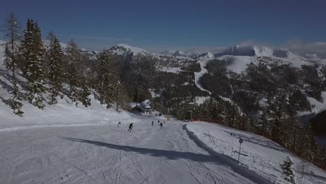 Luftaufnahme-Eines-Skigebiets-An-Einem-Sonnigen-Tag-Mit-Menschen-Auf-Der-Skipiste-Und-Der-Kamera,-Die-Den-Skifahrern-Folgt,-Die-Den-Hang-Hinunterfahren,-Mit-Wunderschönen-Bergen-Im-Hintergrund