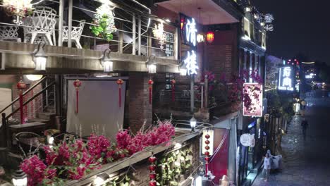 Vuelo-De-Drones-Sobre-Un-Restaurante-Chino-Iluminado-Al-Aire-Libre-En-Un-Callejón-De-La-Ciudad-De-Guangzhou-Donde-Se-Ve-Gente-En-La-Calle