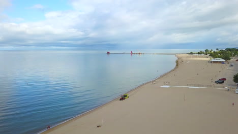 Aerial-drone-shot-over-a-tractor-driving-down-a-large-sandy-beach-with-large-lake-and-a-lighthouse-in-the-background-on-a-cloudy-day