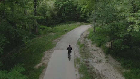 drone-following-man-exercising-by-riding-a-bike-down-a-quiet-bike-path-in-the-woods-wearing-a-black-outfit