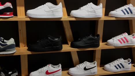 Panning-scene-of-various-assorted-branded-unisex-gender-sneakers---trainers-displayed-on-a-woodern-rack-shelves-with-black-background