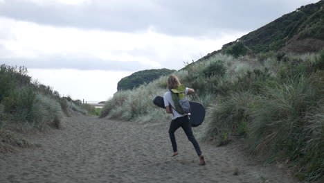 Young-blonde-man-running-happily-towards-the-ocean,-with-a-backpack-and-a-guitar-cover,-on-a-sandy-walkway-surrounded-by-high-grass-at-Bethells-Beach,-on-a-cloudy-and-windy-day