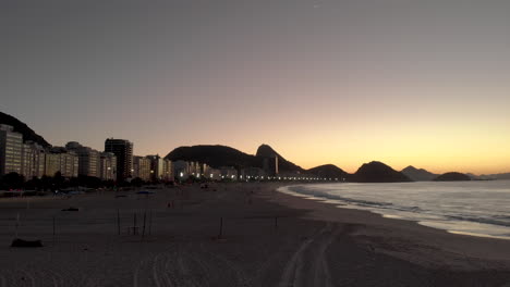Aerial-view-of-Copacabana-beach-early-in-the-morning-before-sunrise-at-golden-hour-with-the-city-lights,-Sugarloaf-mountain-and-hills-of-Niteroi-in-the-background