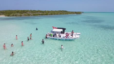 AERIAL-VIEW-PEOPLE-ENJOY-BEACH-PARTY-inside-sea-WATER-with-BOATS-MOORED,-Los-Roques