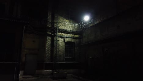 Dark-abandoned-prison-yard,-sharp-fences-visible-at-night,-the-outside-lamp-is-shining