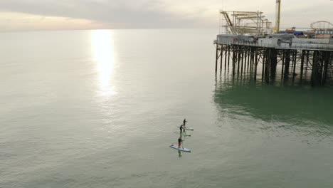 Goldener-Sonnenuntergang-über-Dem-Meer-Mit-Drei-Personen-Auf-Stand-Up-Paddle-Boards,-Die-Am-Brighton-Pier-Vorbeifahren