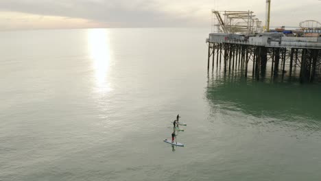 Puesta-De-Sol-Dorada-Sobre-El-Océano-Con-Tres-Personas-En-Tablas-De-Remo-Pasando-Por-El-Muelle-De-Brighton