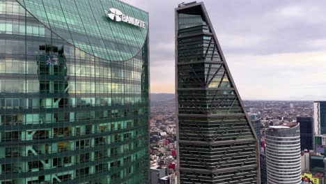 Exhilarating-high-aerial-flight-through-narrow-opening-between-tall-torre-mayor-tower-skyscraper-and-reforma-glass-building-on-cloudy-sky-day-toward-mountain-range,-Mexico-city,-drone-approach