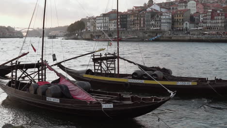 Close-up-view-of-two-rabelo-sailboats-with-port-wine-barrels,-moored-on-the-Douro-River,-with-old-town-Porto-and-Arrabida-bridge-in-the-background