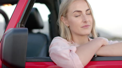 Young-Caucasian-woman-with-blonde-hair-smiles-gently,-leaning-on-a-red-car-door-during-a-road-trip