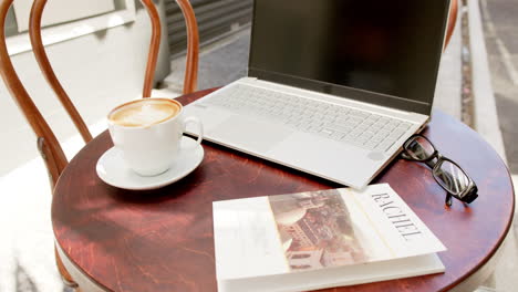 A-laptop,-a-cup-of-coffee,-glasses,-and-a-magazine-rest-on-a-cafe-table