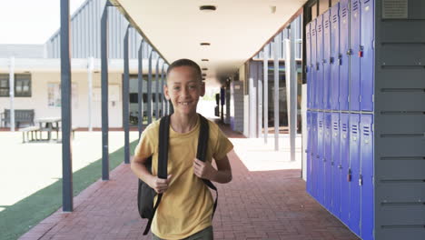 Biracial-boy-with-a-backpack-stands-smiling-in-a-school-corridor