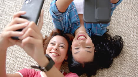 Two-young-biracial-female-friends-are-lying-on-the-floor,-taking-a-selfie-with-their-phones