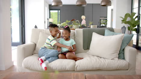 African-American-brother-and-sister-sit-together-on-a-sofa-at-home,-sharing-a-tablet
