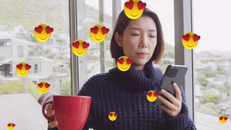 Animation-Von-Emoji-Symbolen-über-Einer-Asiatischen-Frau,-Die-Kaffee-Trinkt-Und-Zu-Hause-Ihr-Smartphone-Benutzt