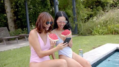 Zwei-Freundinnen-In-Badeanzügen-Genießen-Wassermelone-An-Einem-Pool,-Eine-Zeigt-Etwas-Auf-Einem-Telefon