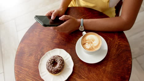 Junge-Frau-Mit-Gemischter-Abstammung,-Die-Ein-Smartphone-An-Einem-Cafétisch-Mit-Einem-Donut-Und-Kaffee-In-Einem-Café-Benutzt