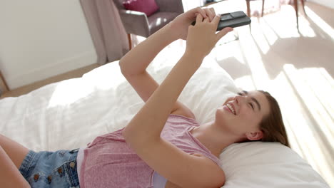 Una-Adolescente-Caucásica-Con-Cabello-Castaño-Usa-Un-Teléfono-Inteligente-Mientras-Está-Acostada-En-Una-Cama-En-Casa