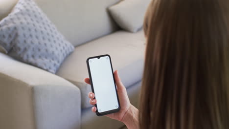 A-Caucasian-woman-is-holding-a-smartphone-with-a-blank-screen-on-the-couch-at-home,-copy-space