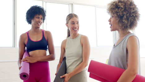 Three-women-are-chatting-in-bright-yoga-studio,-holding-mats